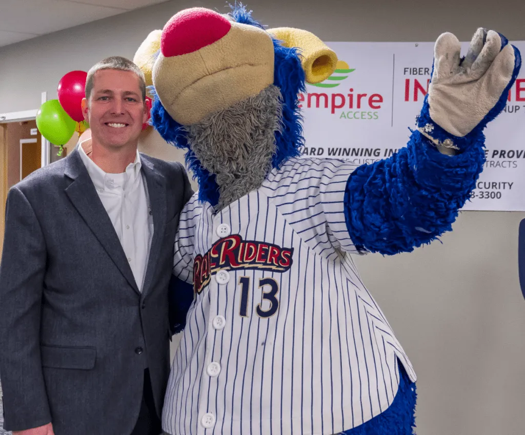 A man in a suit stands smiling next to a person in a blue and yellow bear mascot costume wearing a pinstriped jersey with the number 13. They are indoors, with colorful balloons and a business sign in the background.