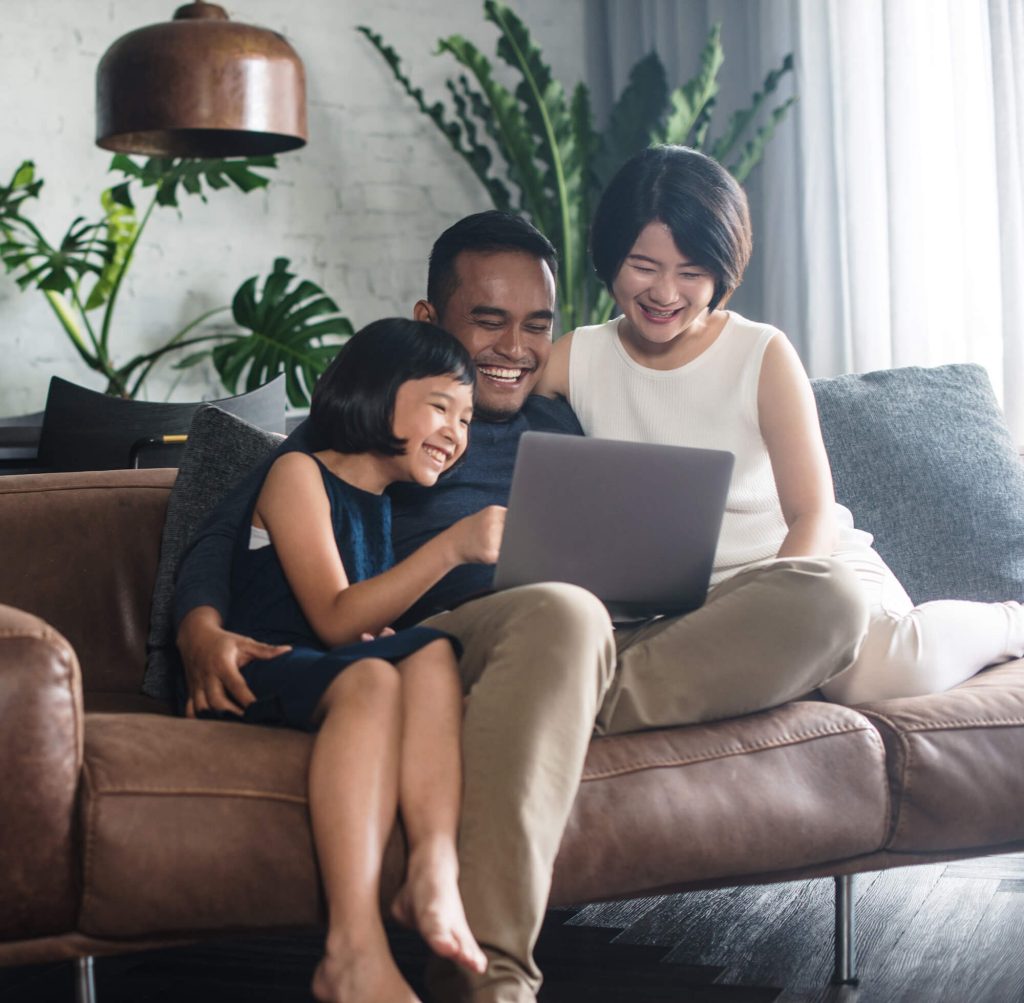 A family of three sits on a couch, smiling and looking at a laptop, enjoying the benefits of high-speed internet. A man sits in the middle, with a woman on his right and a young girl on his left. They appear happy and relaxed in a cozy living room with plants in the background.