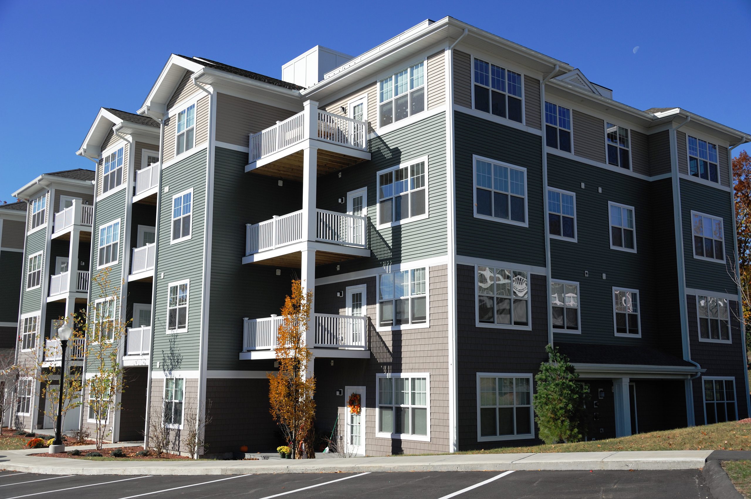 A modern four-story MDU with green and beige siding, featuring multiple balconies. The building is surrounded by small trees beneath a clear blue sky. A parking lot is visible in the foreground, offering ample space for residents.