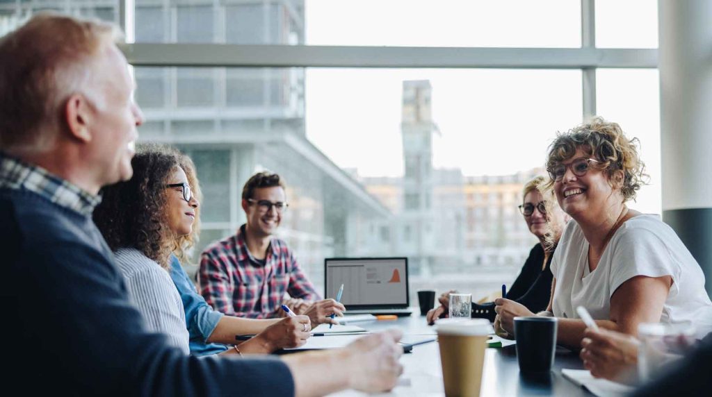 A group of six people sitting around a table in a modern office, engaged in a meeting about services. Laptops and papers are spread across the table, and large windows in the background reveal an urban cityscape. Everyone appears to be smiling and interacting.