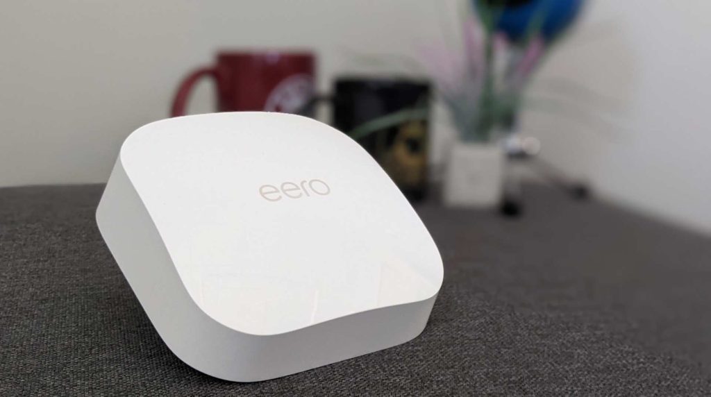 A white Eero mesh Wi-Fi router sits on a gray surface. In the blurred background two mugs are visible.