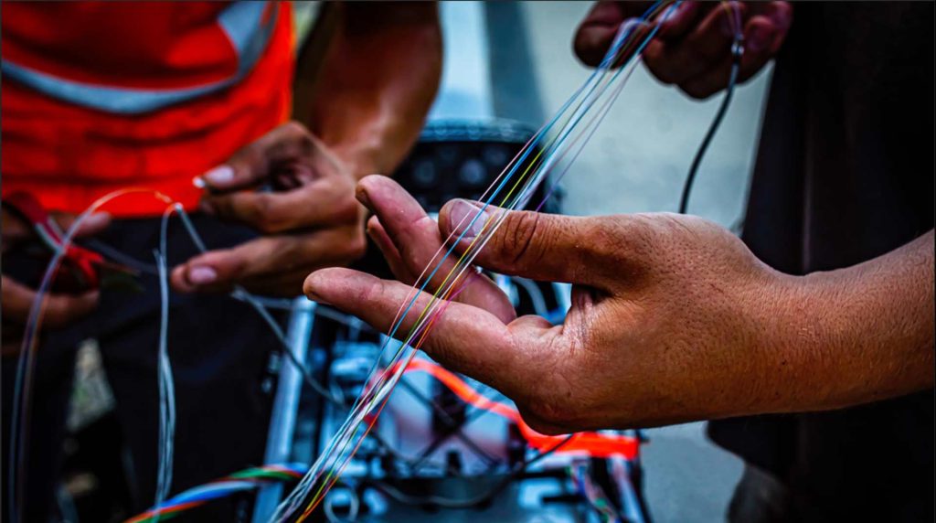 Close-up of two people working with fiber optic cables. One person holds several colorful strands while another uses tools on nearby equipment. The scene suggests the installation or repair of advanced fiber optic telecommunications infrastructure.