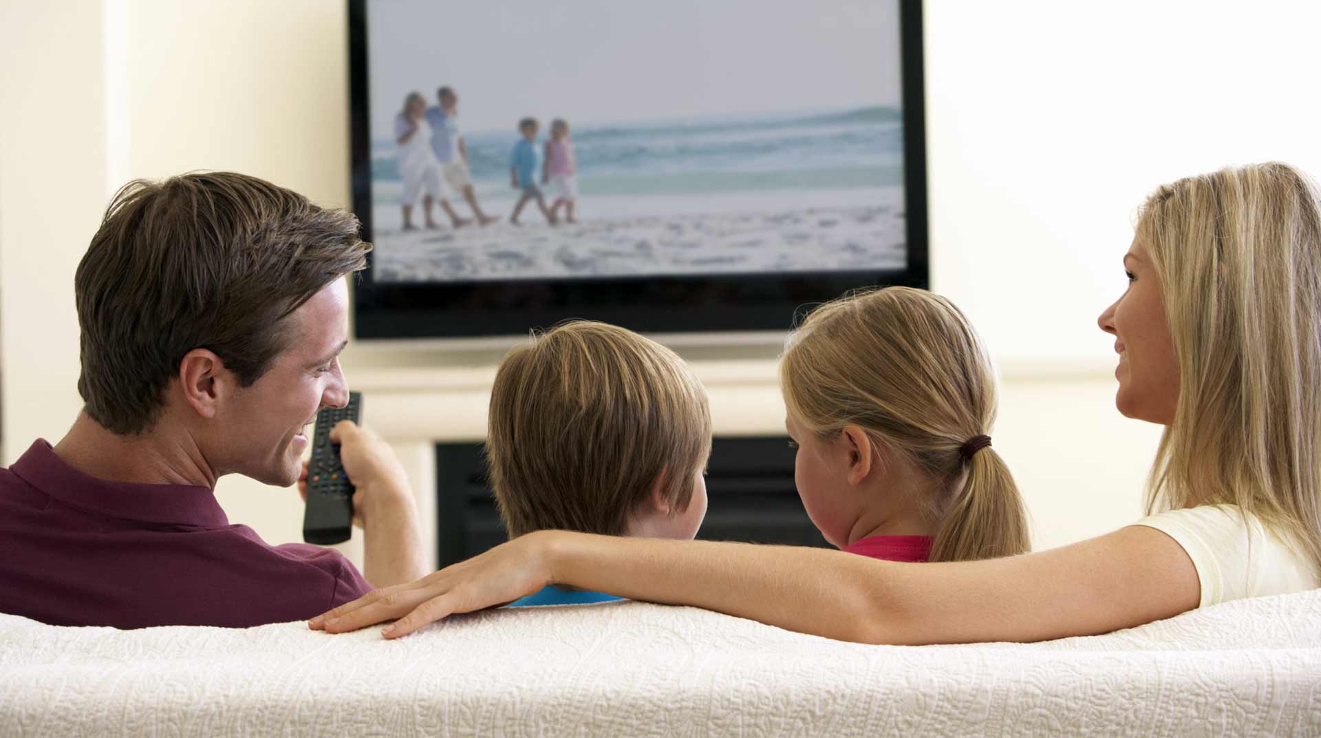 A family of four, sitting on a sofa, enjoys the right TV option for their cozy evening together. The man holds the remote control with a sense of satisfaction as the screen reveals a serene beach scene with people strolling along the shore, creating a relaxed atmosphere.