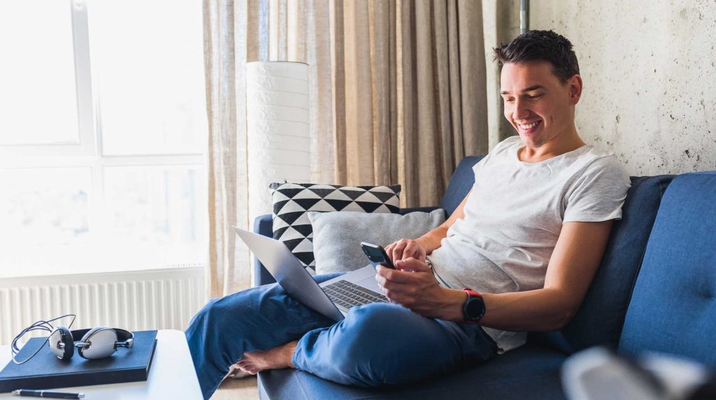 Man sitting on his couch looking at his laptop with a tablet and headphones sitting on his table