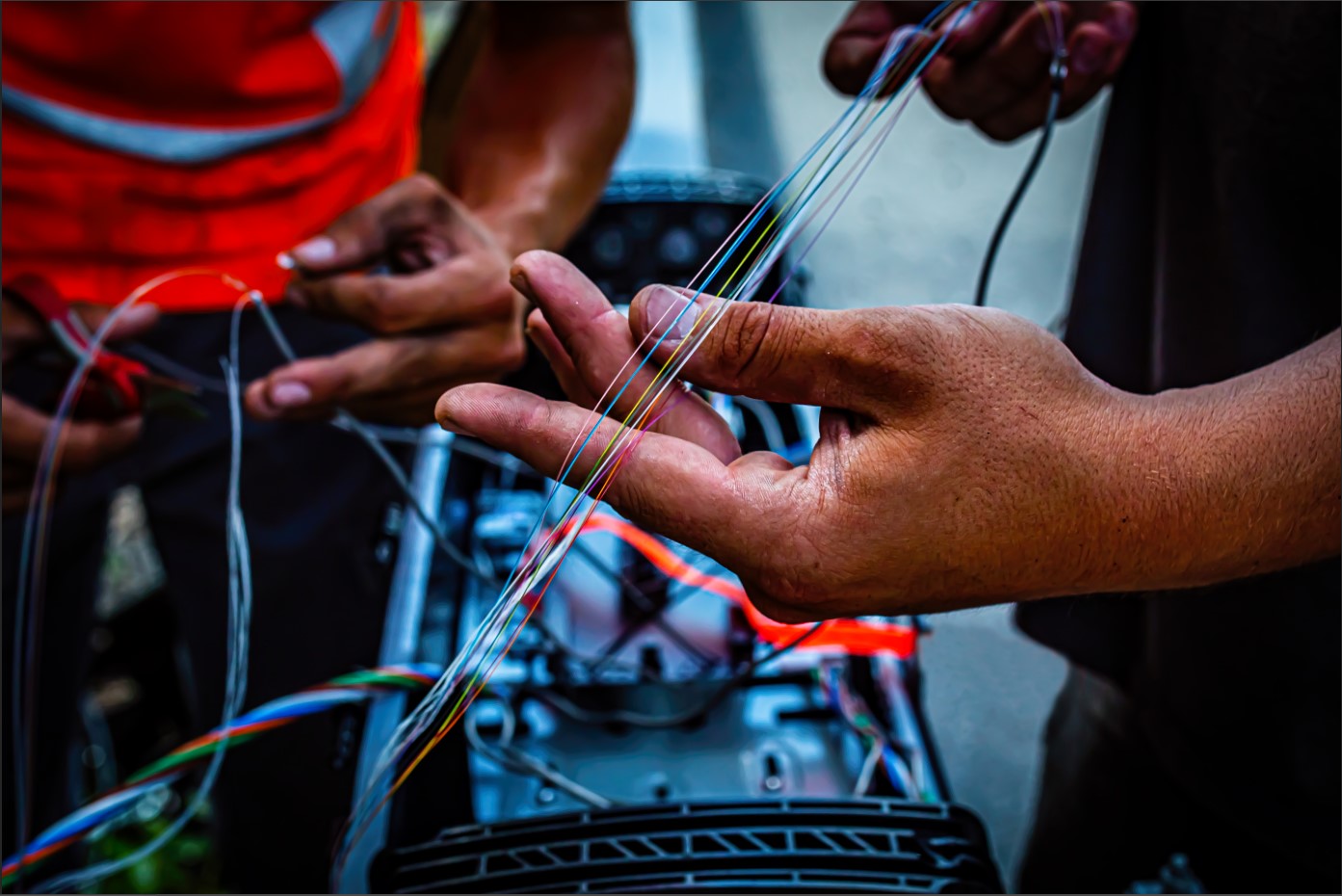 Close-up of two people working with fiber optic cables. One person holds several colorful strands while another uses tools on nearby equipment. The scene suggests the installation or repair of advanced fiber optic telecommunications infrastructure.