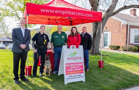 A group of five people and a police dog stand smiling under a red tent with "empireaccess.com" on it, celebrating Empire Access's latest fiber expansion. A sign in front advertises their services. They are outdoors in front of a brick building.