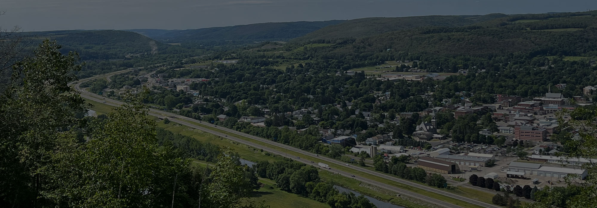Aerial view of a small town surrounded by lush green hills and forests. A highway runs parallel to a river, with residential and commercial buildings scattered throughout the landscape under a clear blue sky, all connected seamlessly through high-speed internet for Bath residents.