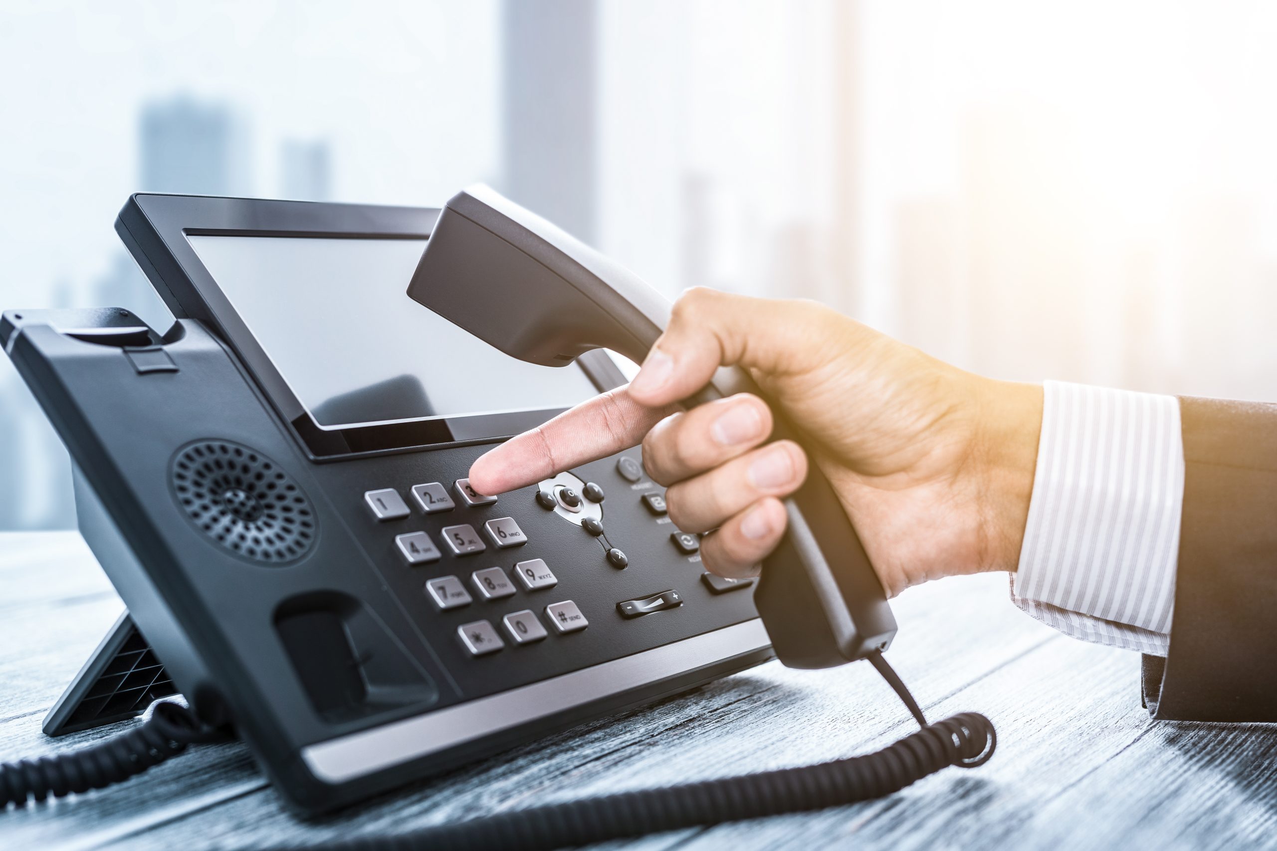 A person in a suit is using the Empire Phone Service, holding the receiver of a black office phone while pressing a button on the keypad. The phone sits on a desk against a bright, blurred office background, exemplifying streamlined communication.