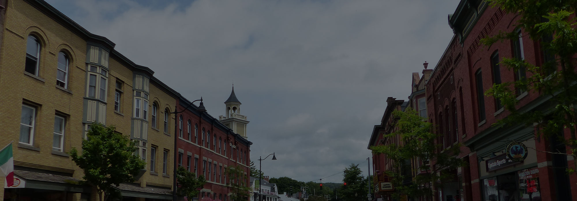 A quaint small town street with historic brick buildings lines both sides, adding charm to Hamilton. A few trees provide a touch of greenery, and a clock tower rises in the background under a partly cloudy sky—a blend of timeless elegance in the digital age.