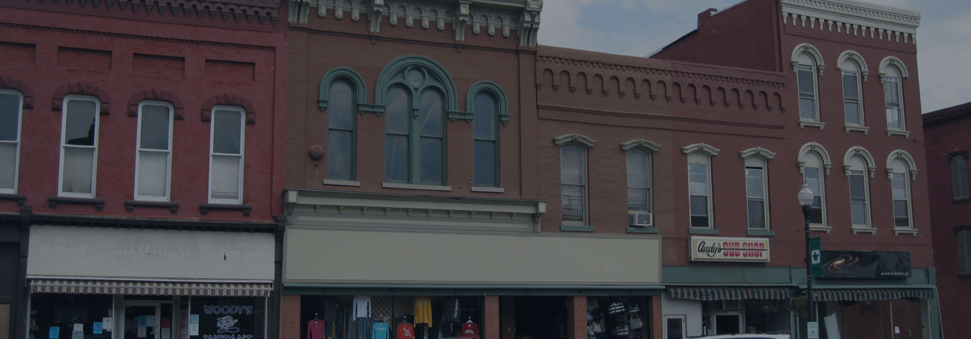Street view of a row of historic brick buildings with colorful facades in Hornell. The scene includes a clothing store showcasing shirts and a restaurant with a sign featuring bold red letters. A street lamp stands prominently in front, adding to the charming ambiance both offline and on the internet.