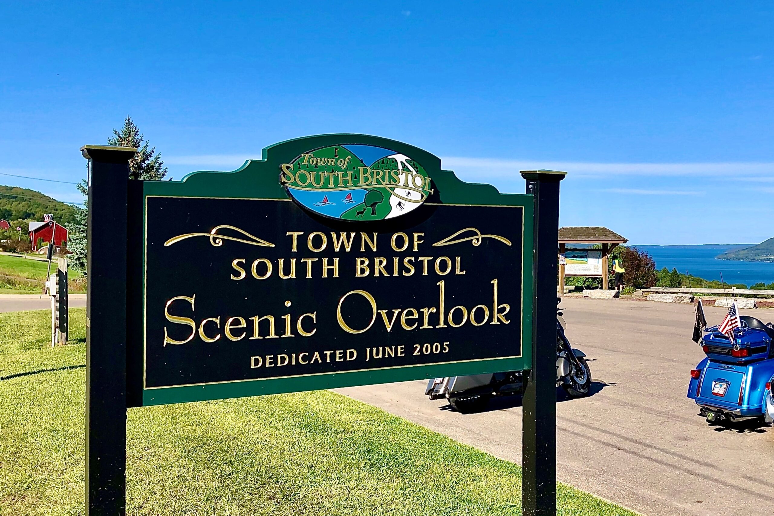 A sign reading "Town of South Bristol Scenic Overlook, Dedicated June 2005," stands proudly against a backdrop of rolling green hills, clear blue skies, and a shimmering glimpse of water—a testament to the town's empire expansion in embracing nature's magnificence.