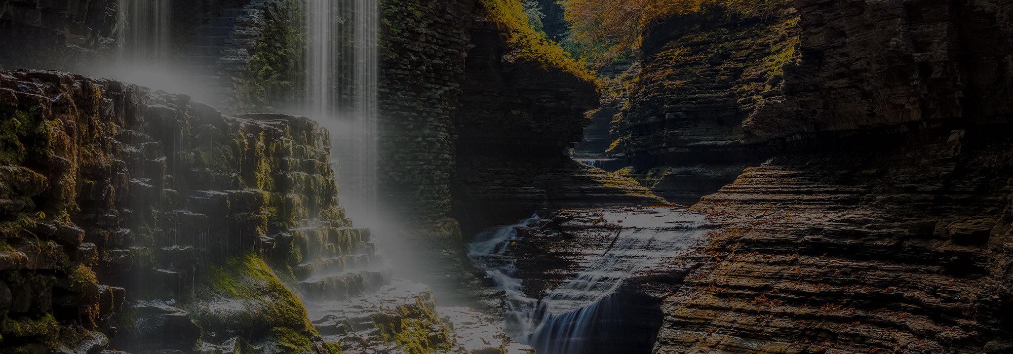 A serene waterfall cascades over moss-covered stone ledges into a rocky gorge at Watkins Glen. Sunlight filters through gaps in the trees, illuminating the mist and creating a peaceful atmosphere, as if untouched by the hustle of the internet age.