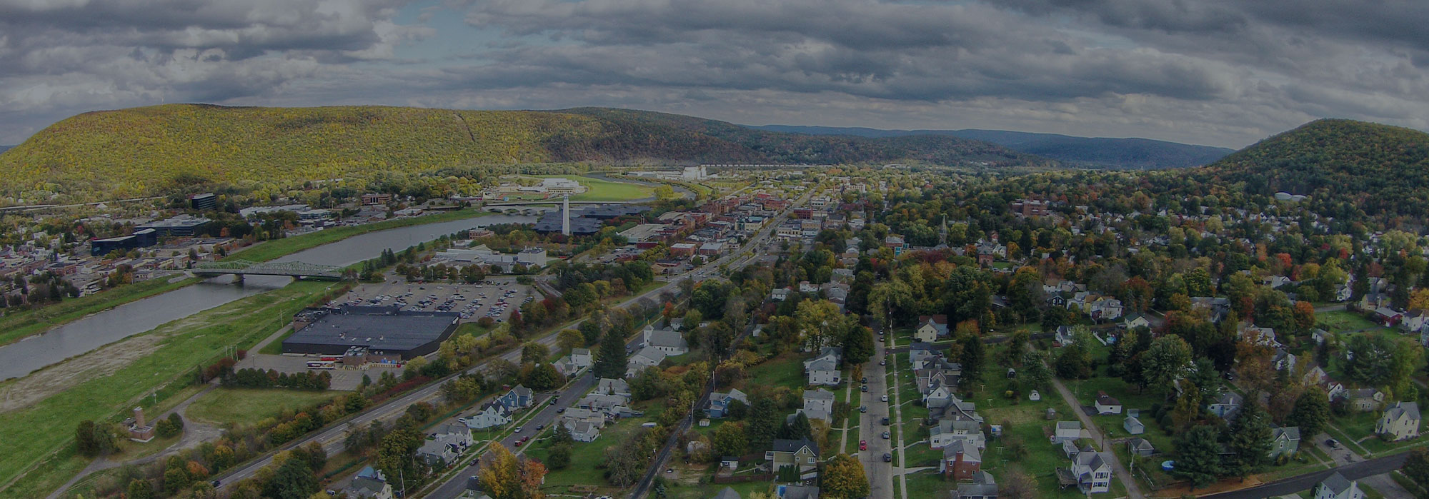 Aerial view of a small town with scattered houses, a river, and a bridge. The surrounding landscape features lush greenery and hills under a cloudy sky, all seamlessly connected like Corning Internet’s network weaving through the community.