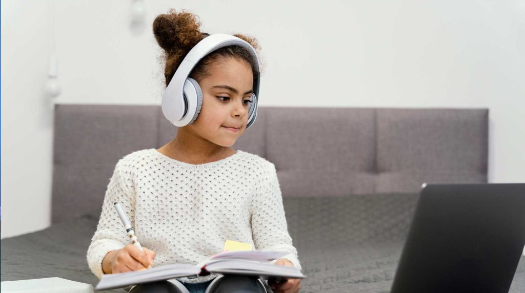 Young girl sitting on her couch with a notebook and pencil in her hand, doing her home work by listening with headphones to her laptop