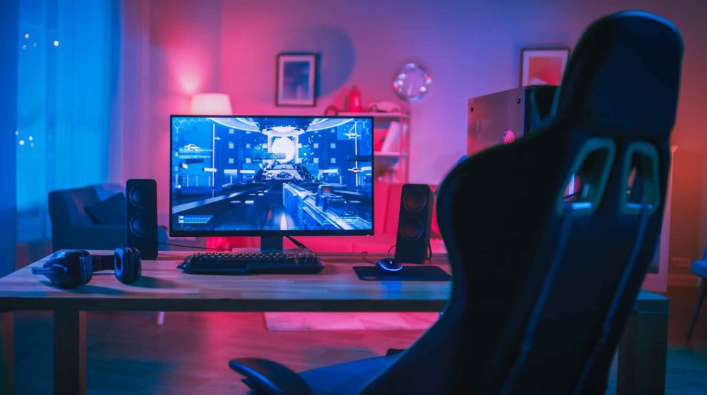 A gaming setup with a computer monitor displaying a futuristic game immerses you in an electrifying atmosphere. The room glows with blue and pink ambient lighting, while a gaming chair, keyboard, speakers, and headphones sit on the wooden desk. Cozy interior touches include a sofa and framed pictures in the background.
