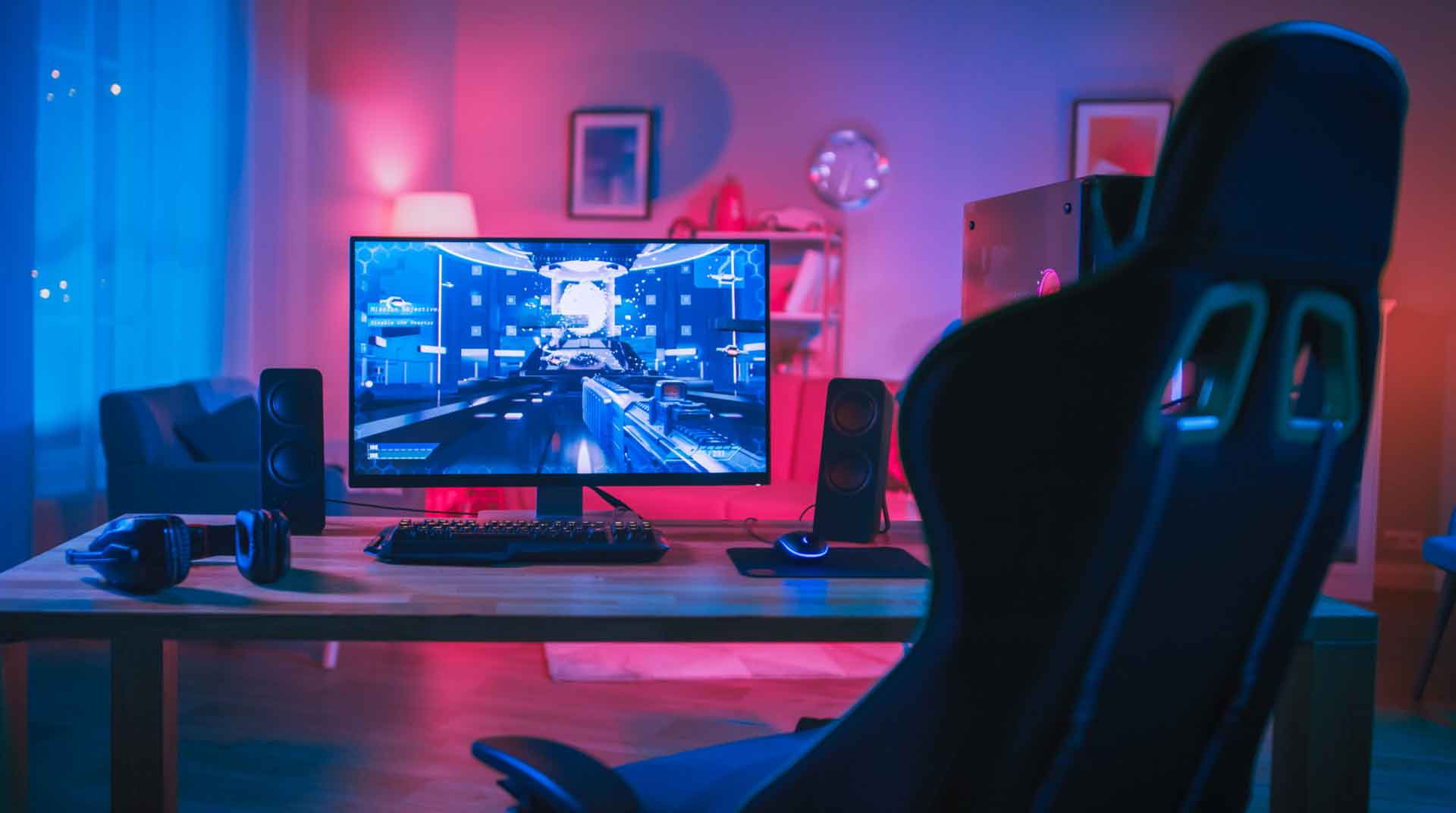 A gaming setup with a computer monitor displaying a futuristic game immerses you in an electrifying atmosphere. The room glows with blue and pink ambient lighting, while a gaming chair, keyboard, speakers, and headphones sit on the wooden desk. Cozy interior touches include a sofa and framed pictures in the background.