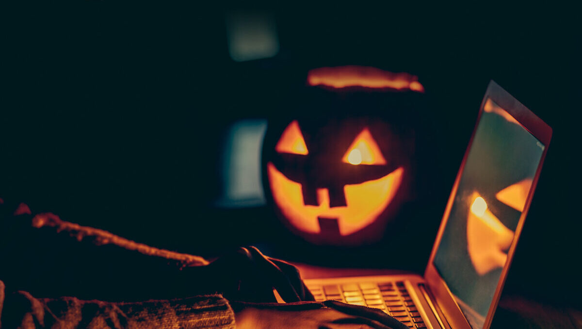 A person types on a laptop in a dimly lit room, with a glowing jack-o'-lantern and fiber optic decorations casting an eerie light. The atmosphere is dark and mysterious, capturing a Halloween theme.