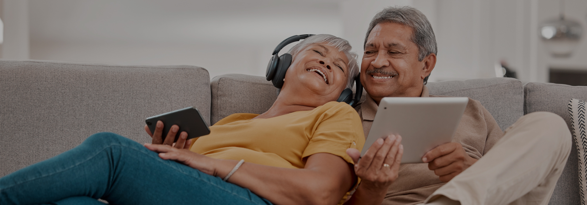 An older couple relaxes on a couch. The woman, wearing headphones, leans against the man and holds a smartphone, smiling. The man, smiling, holds a tablet as they enjoy the most reliable fiber internet in the Northeast. Both appear content and comfortable in a bright living room.