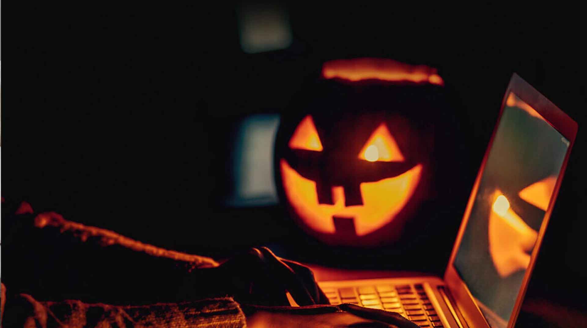 A person types on a laptop in a dimly lit room, with a glowing jack-o'-lantern and fiber optic decorations casting an eerie light. The atmosphere is dark and mysterious, capturing a Halloween theme.