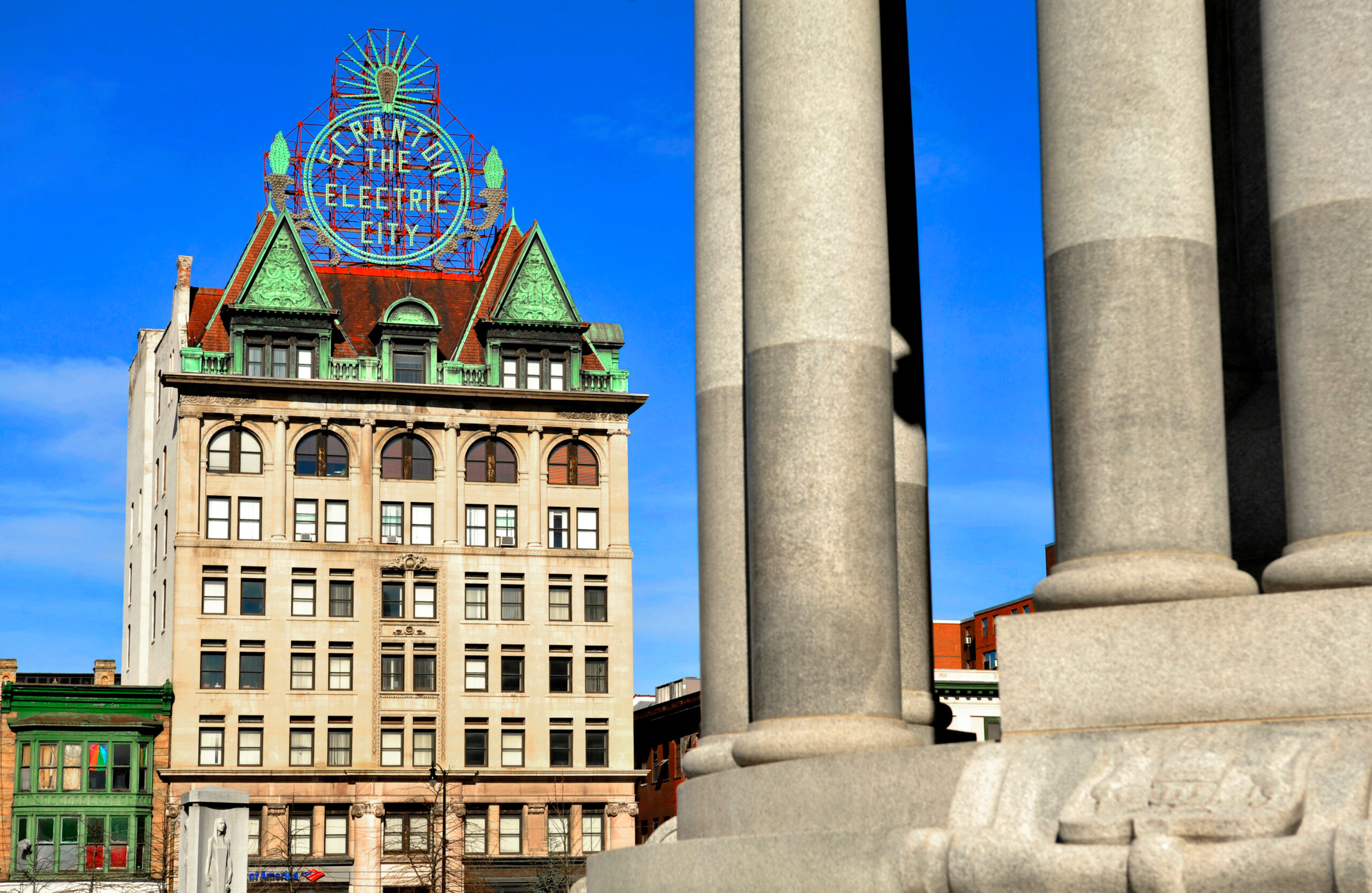 A historic building in Scranton, featuring a green roof and red accents, proudly displays a neon sign proclaiming "Scranton The Electric City" against the vibrant blue sky. In the foreground, stone columns add to the city's timeless charm.