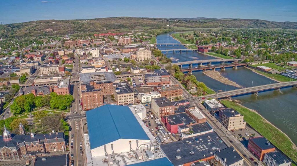 Aerial view of a city in the Twin Tiers, with multiple bridges crossing a river. The cityscape nestles among green hills under a clear blue sky. Buildings of various sizes populate the area, with a prominent white building in the foreground.