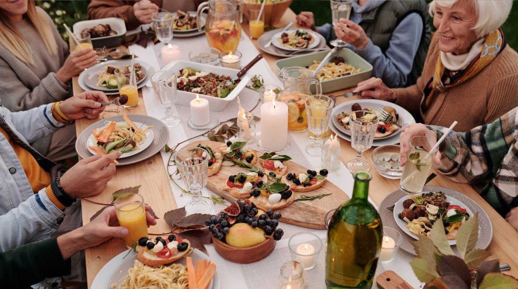 A group of people giving thanks while enjoying a meal around a wooden table set with various dishes, fruits, and drinks. Candles are lit, creating a cozy atmosphere. The diners are smiling and engaged in conversation, surrounded by greenery.