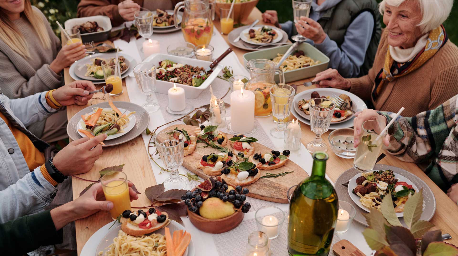 A group of people giving thanks while enjoying a meal around a wooden table set with various dishes, fruits, and drinks. Candles are lit, creating a cozy atmosphere. The diners are smiling and engaged in conversation, surrounded by greenery.