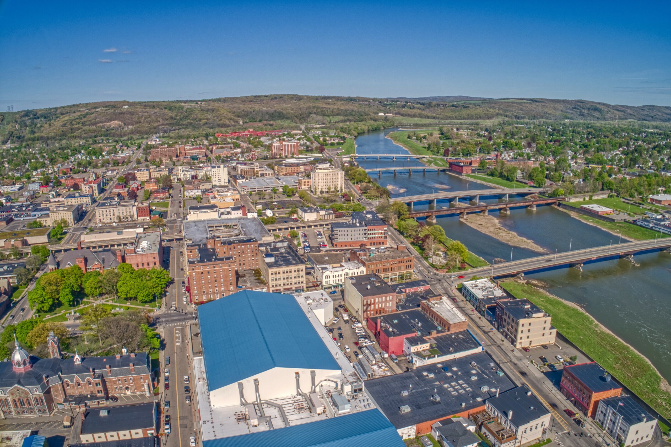 Aerial view of a city in the Twin Tiers, with multiple bridges crossing a river. The cityscape nestles among green hills under a clear blue sky. Buildings of various sizes populate the area, with a prominent white building in the foreground.