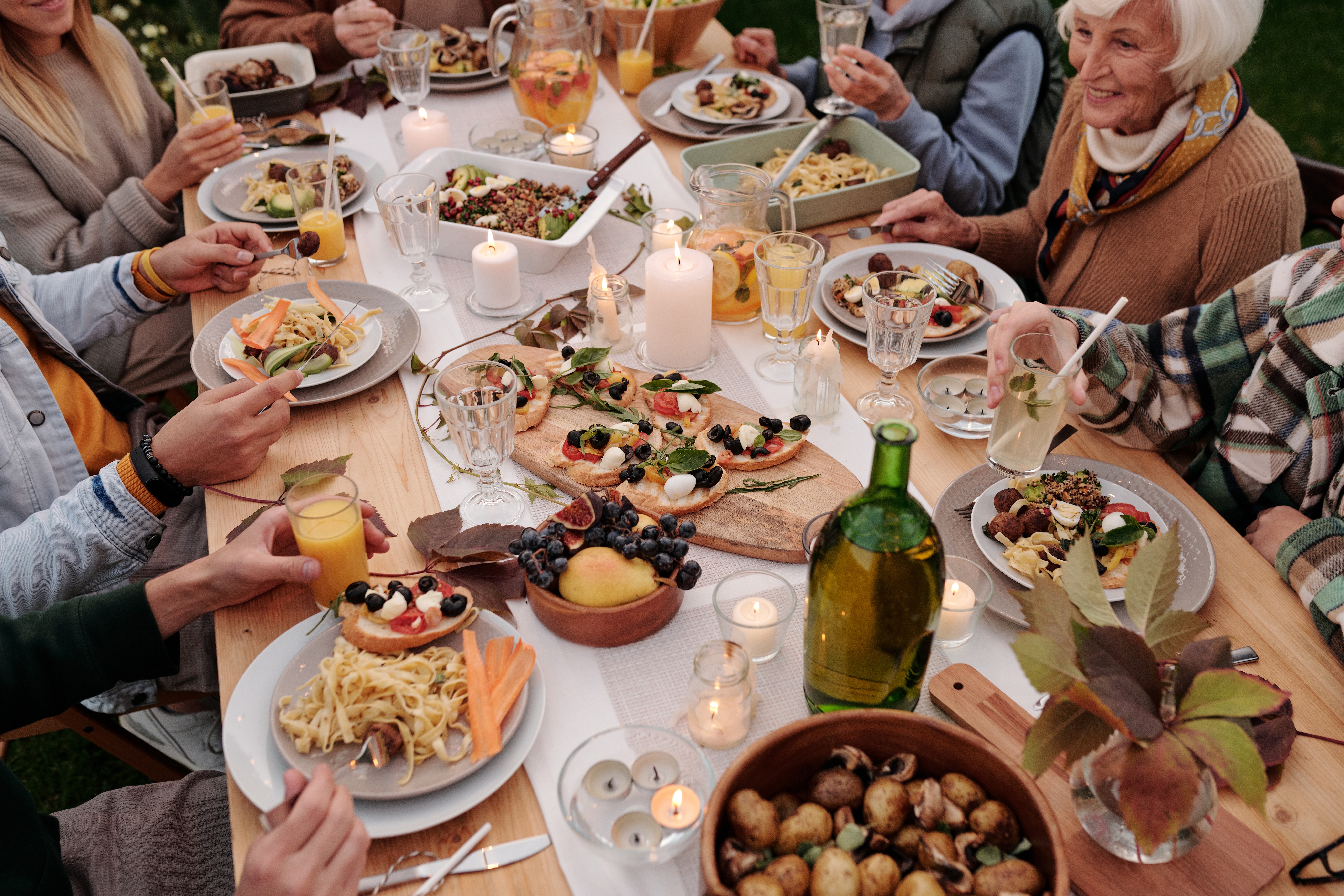 A group of people giving thanks while enjoying a meal around a wooden table set with various dishes, fruits, and drinks. Candles are lit, creating a cozy atmosphere. The diners are smiling and engaged in conversation, surrounded by greenery.