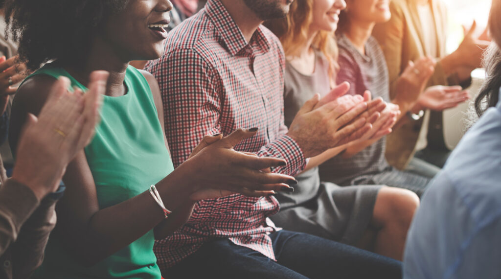 A diverse group of people sitting closely together, smiling and clapping as awards are announced. They are dressed in casual clothing, with sunlight streaming in from the side, creating a warm and cheerful atmosphere.