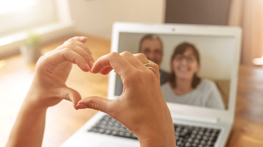 A person forms a heart shape with their hands in front of a laptop screen, where a smiling man and woman chat on a video call. The warm natural light from the window creates an inviting atmosphere, adding purpose to this touching moment of virtual connection.
