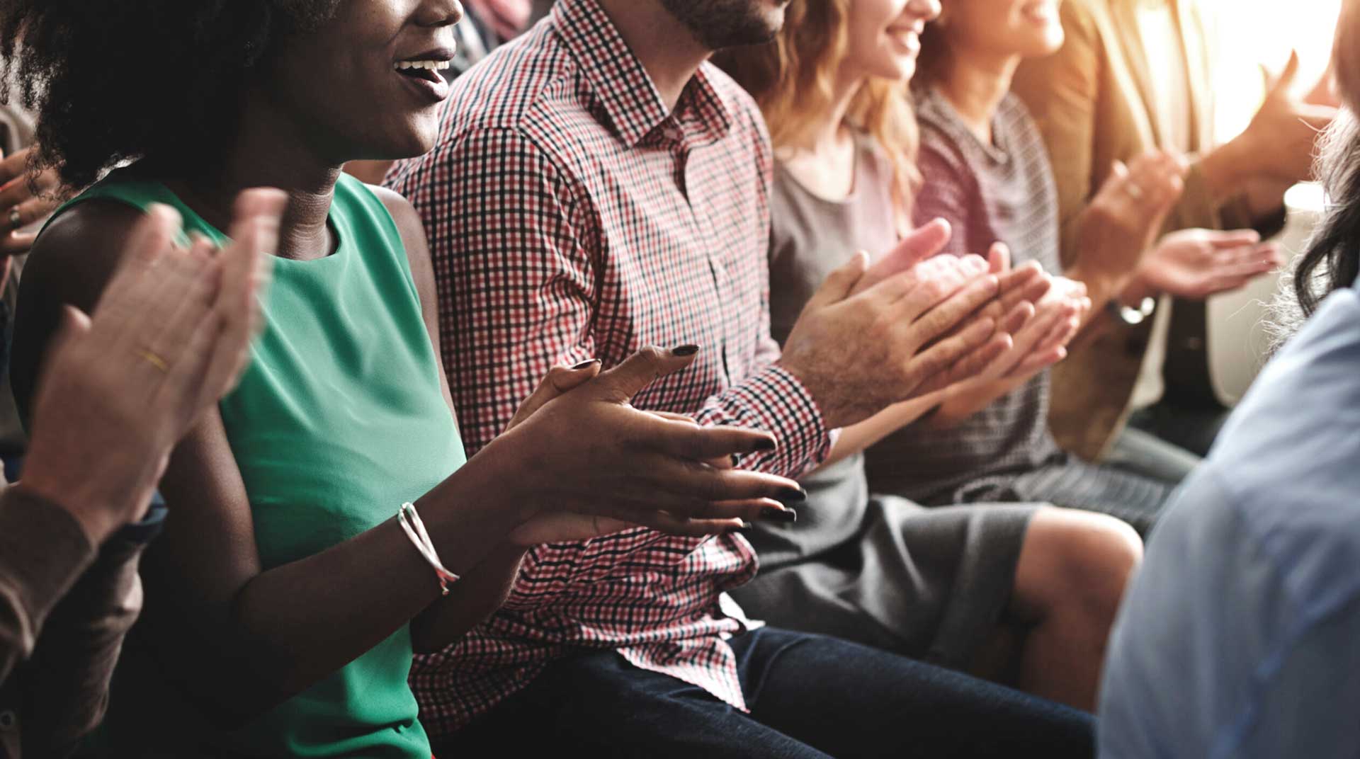 A diverse group of people sitting closely together, smiling and clapping as awards are announced. They are dressed in casual clothing, with sunlight streaming in from the side, creating a warm and cheerful atmosphere.