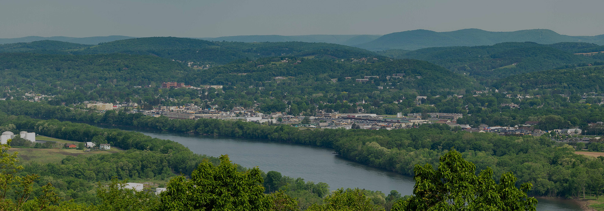 A scenic view of a river meandering through a green, hilly landscape with a town nestled along its banks, reminiscent of picturesque Rochester. Lush forests cover the hills, and the overcast sky casts a serene atmosphere over the valley, much like finding tranquility in your digital escape online.