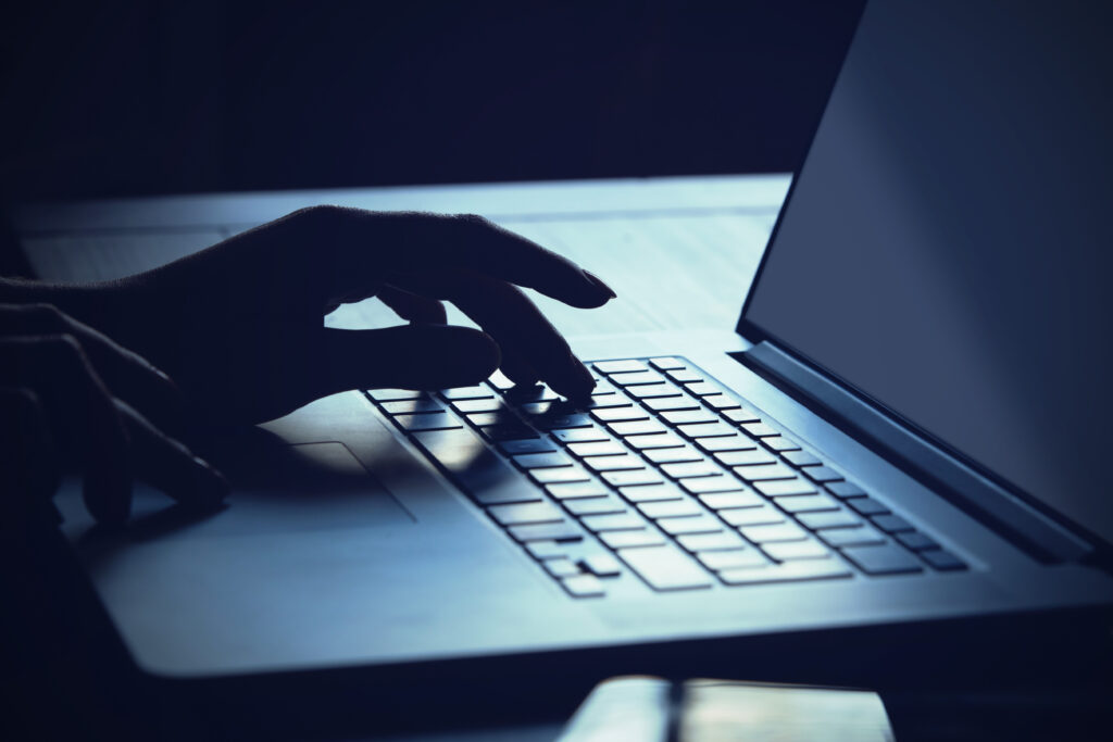In a dimly lit setting, a hand types on a laptop keyboard with symmetrical speed. The screen glows softly, creating a contrast with the dark surroundings. The atmosphere suggests focus and intense concentration on the task at hand.