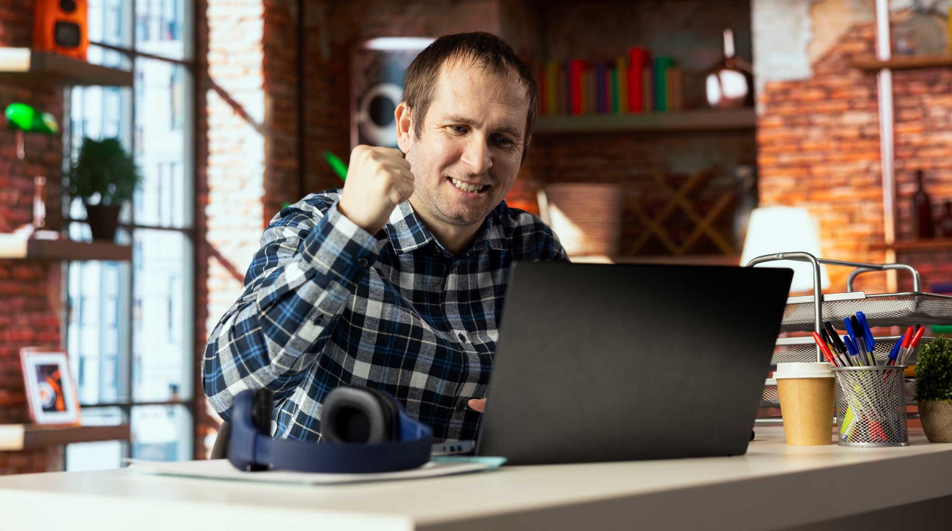 Man sitting with his laptop at a table, smiling with his hand in the air demonstrating excitement