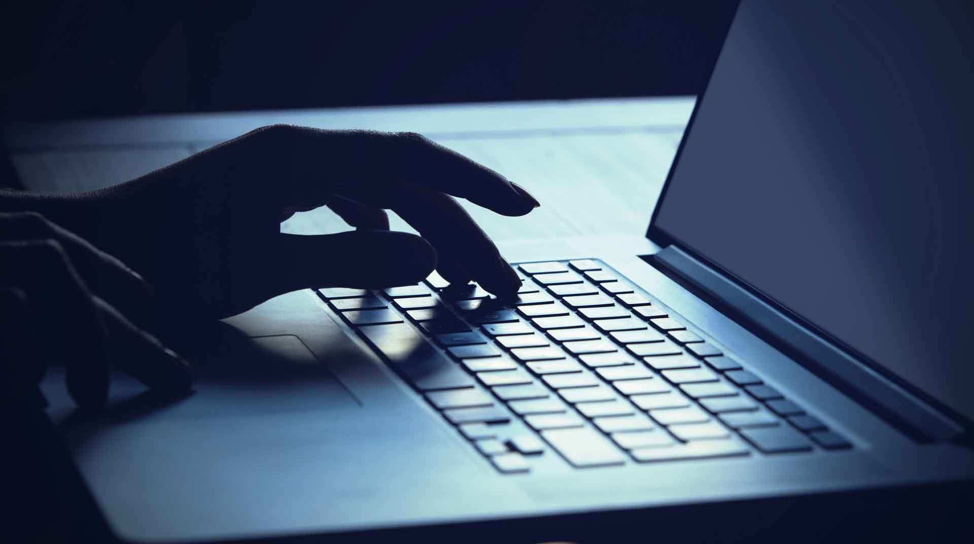 In a dimly lit setting, a hand types on a laptop keyboard with symmetrical speed. The screen glows softly, creating a contrast with the dark surroundings. The atmosphere suggests focus and intense concentration on the task at hand.