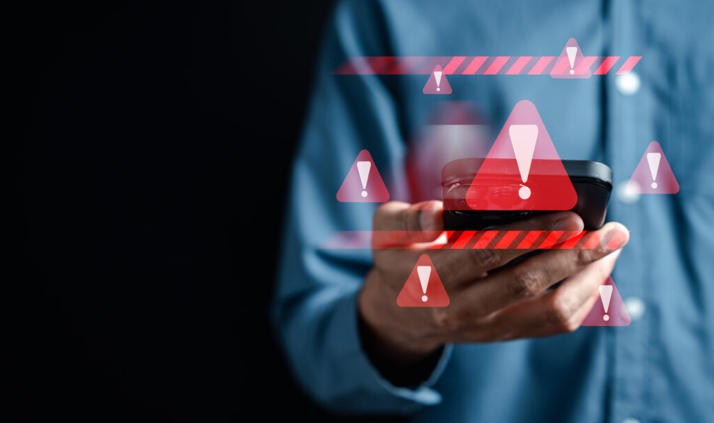 A person in a blue shirt holds a smartphone against a dark background, surrounded by red warning symbols and exclamation marks—hinting at an urgent phone scam alert on their device.