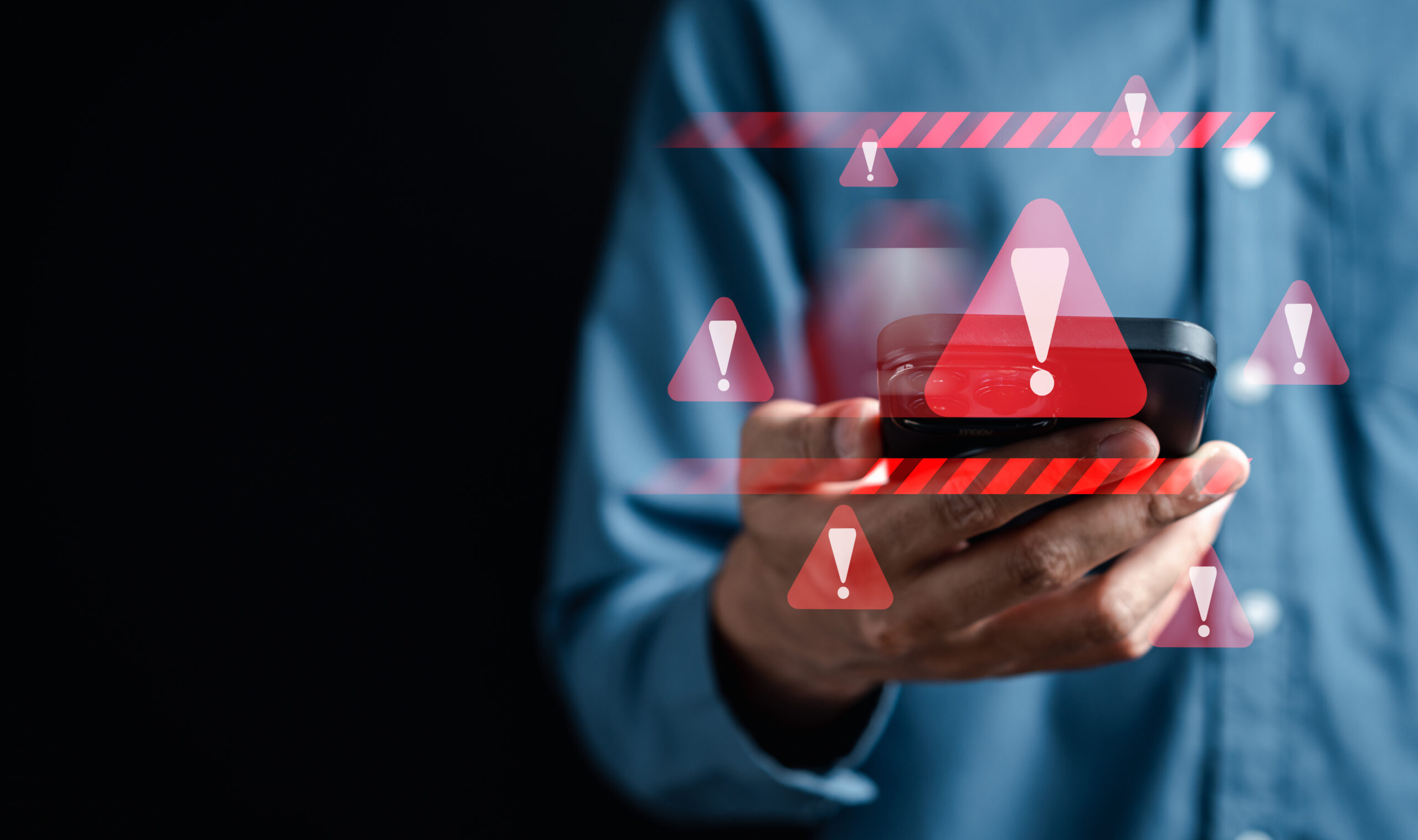 A person in a blue shirt holds a smartphone against a dark background, surrounded by red warning symbols and exclamation marks—hinting at an urgent phone scam alert on their device.