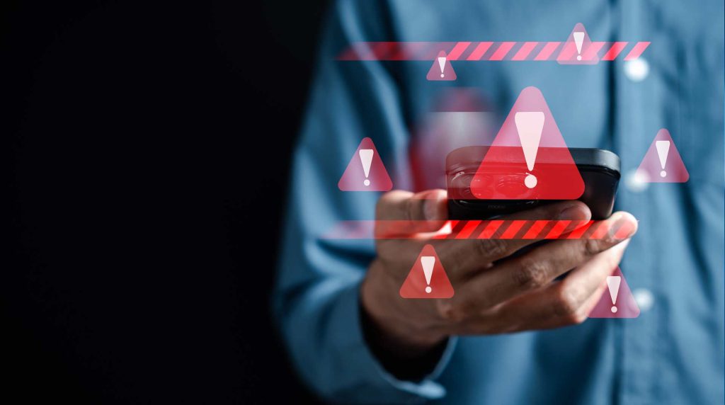 A person in a blue shirt holds a smartphone against a dark background, surrounded by red warning symbols and exclamation marks—hinting at an urgent phone scam alert on their device.