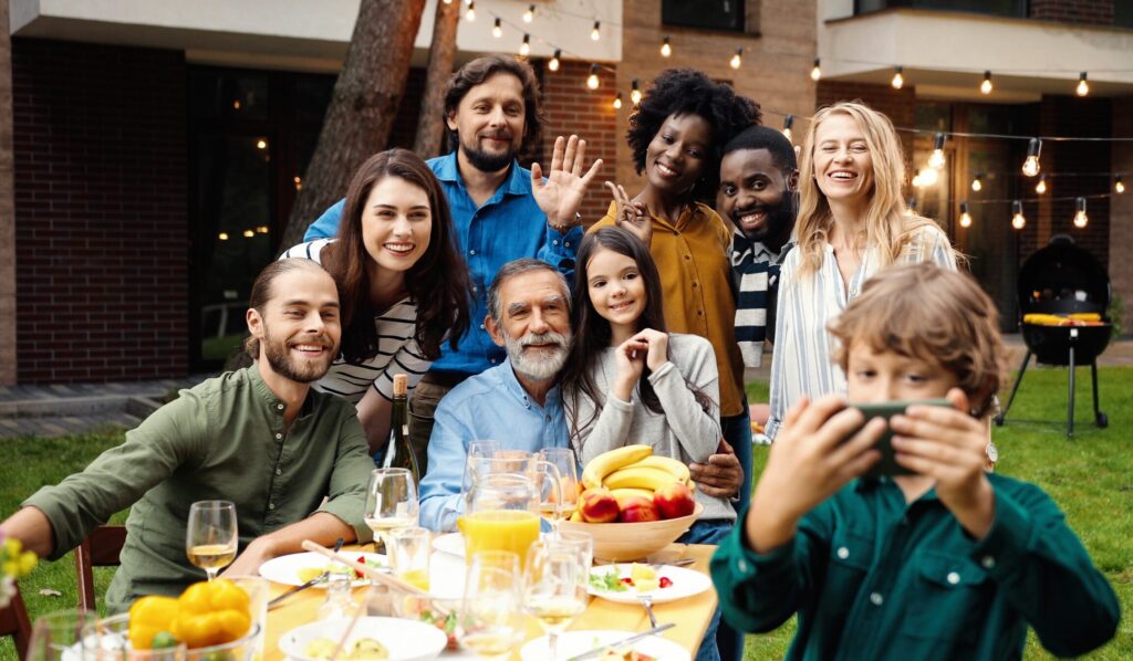 A diverse group of people smiling and posing for a selfie at an outdoor gathering, celebrating their connection. They are seated around a table with food and drinks, while festive string lights twinkle in the background.