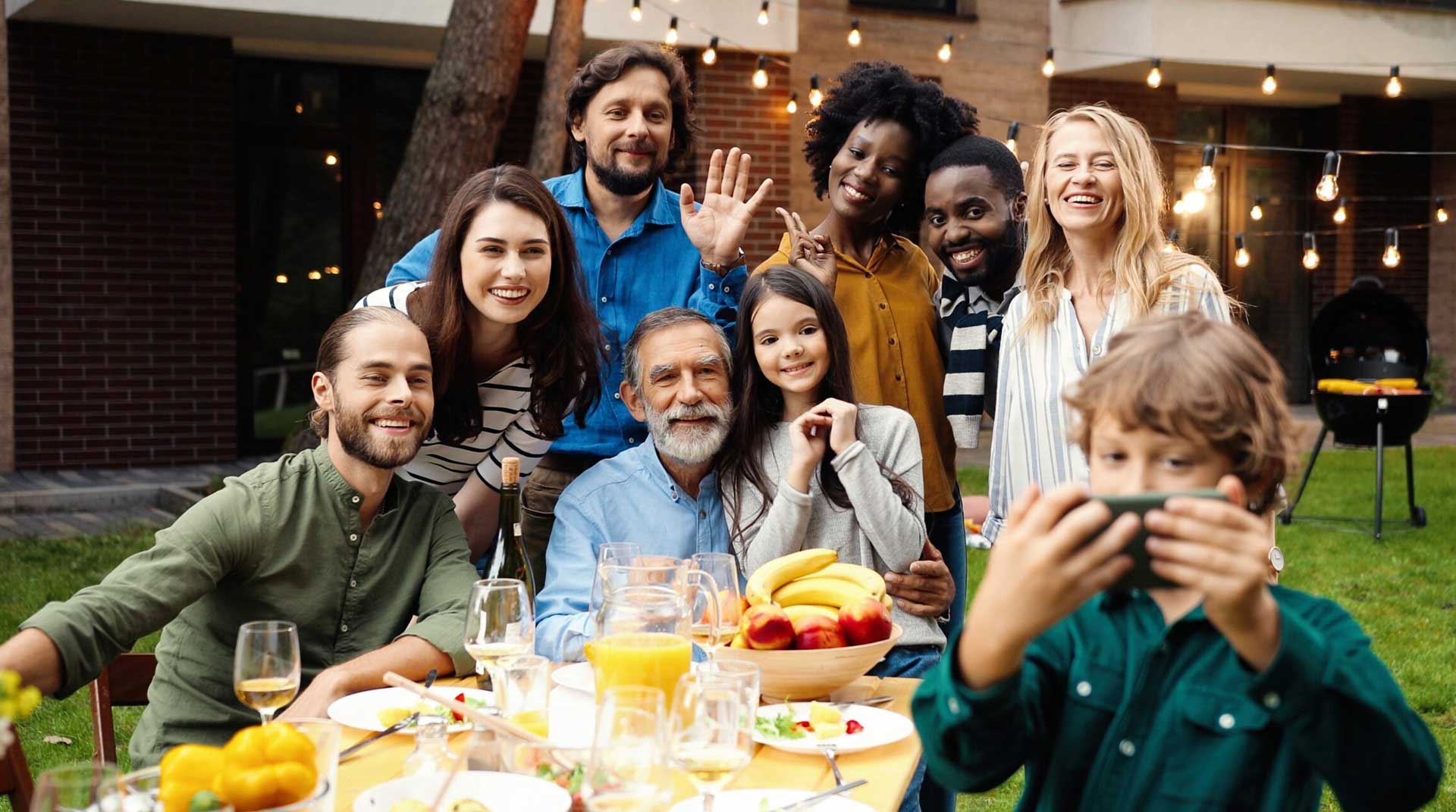 A diverse group of people smiling and posing for a selfie at an outdoor gathering, celebrating their connection. They are seated around a table with food and drinks, while festive string lights twinkle in the background.
