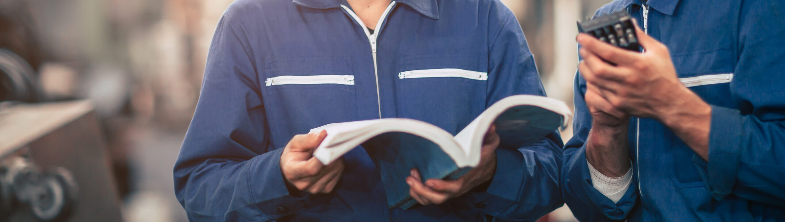 Two people in blue coveralls are diligently working together, one poring over an open book of instructions while the other uses a calculator. They appear to be in an industrial or workshop setting, following guides as they focus intently on their task.
