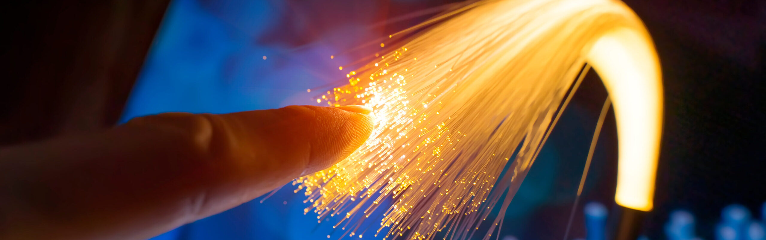 A finger gently touches the dark fiber optic strands, creating a stream of vibrant yellow and orange light against a deep blue background.