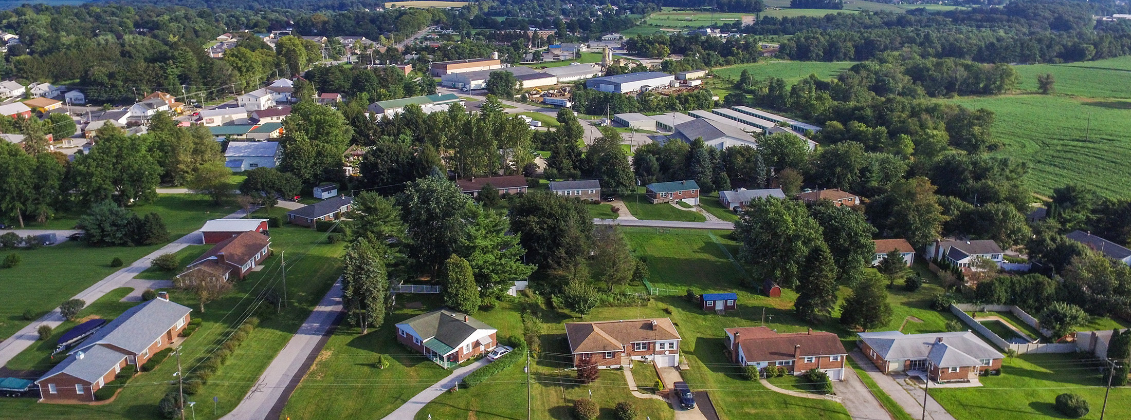 An aerial view showcases the charming neighborhood of a small town, likely Bloomsburg, with houses cradled by lush greenery and trees. Open fields stretch towards a distant cluster of buildings under a clear sky, perhaps reflecting the seamless connectivity of local internet services.