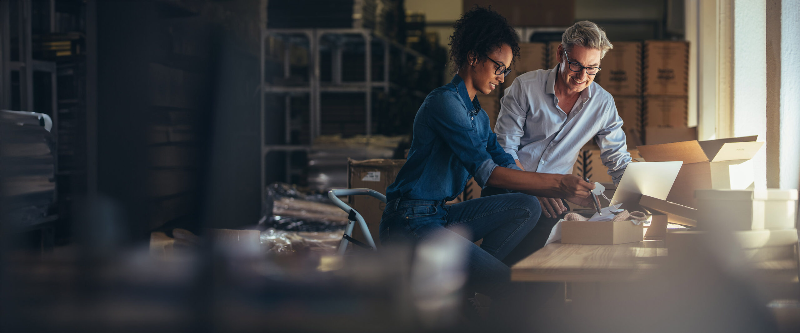 A man and woman working together at a table in a warehouse. The woman, on the left, is writing on a notepad, and the man, on the right, is holding a box while looking at a laptop. They are both smiling and appear to be collaborating.