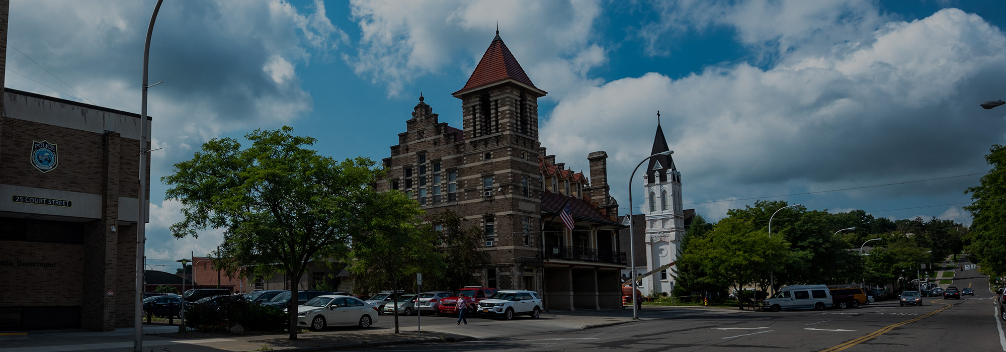 A street scene in Rochester showcases a historic building with a red roof and tower on the left, while a white church with a tall steeple stands on the right. Cars line the road under a cloudy blue sky, blending history with modern elements like internet connectivity.