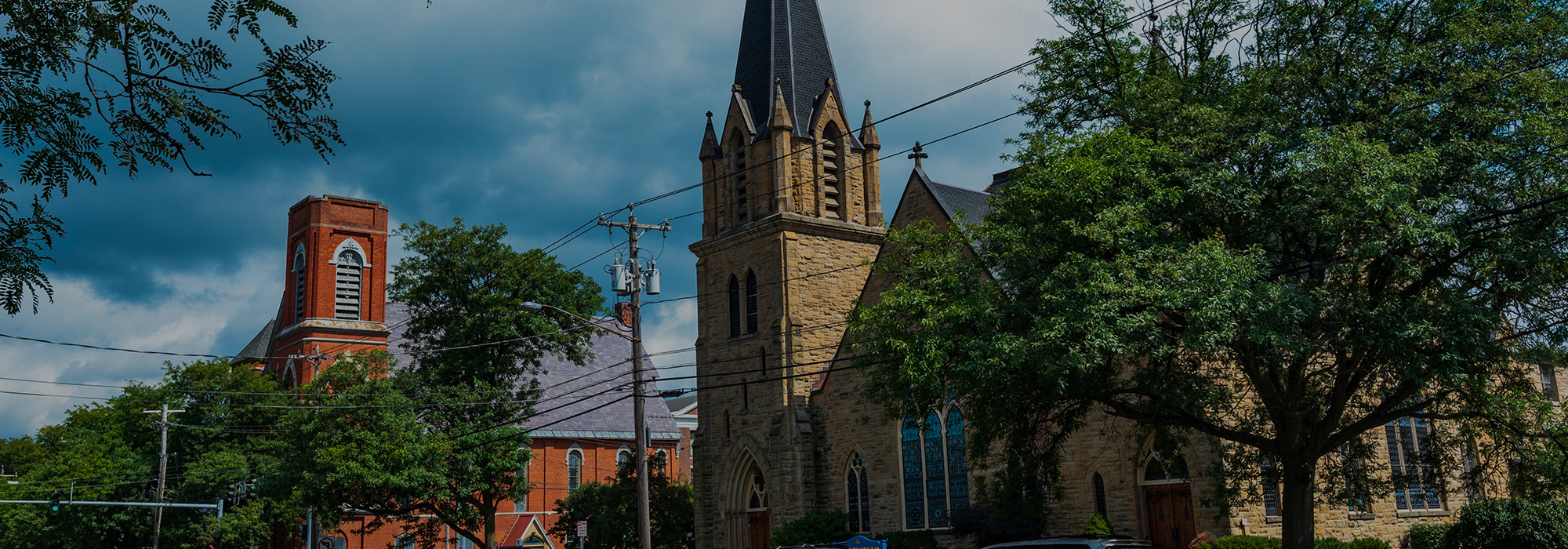 Historic stone church with a tall steeple among lush trees and power lines, capturing the charm of Rochester. A red brick building stands nearby under a cloudy sky, while cars are partially visible on the road, adding life to this picturesque scene in this well-connected Internet age.