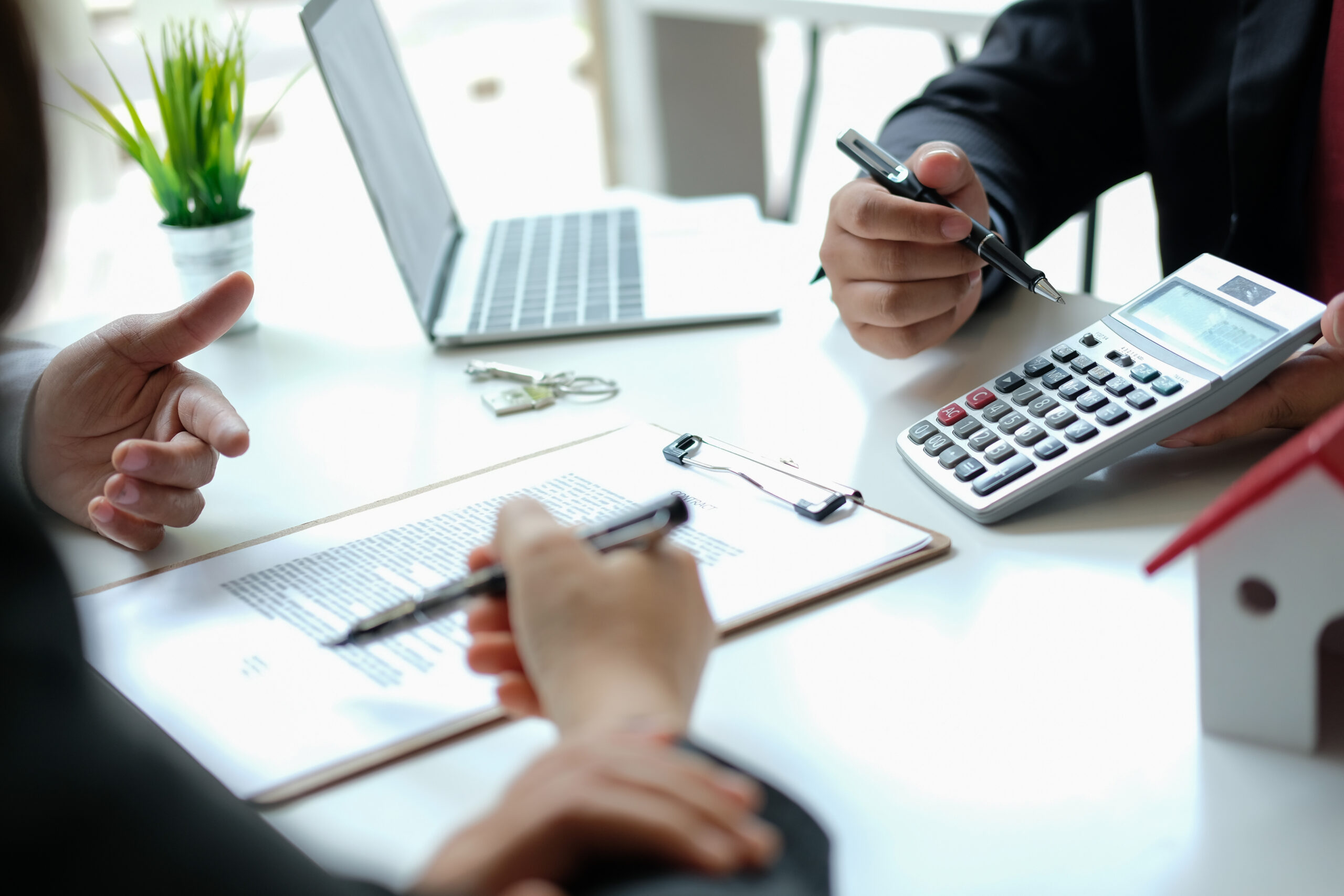 Two people in business attire discuss over a table with documents, a calculator, and pens. In the background, there's a laptop and a small plant. One person gestures with a pen, skillfully accessing finances as they navigate their financial empire.