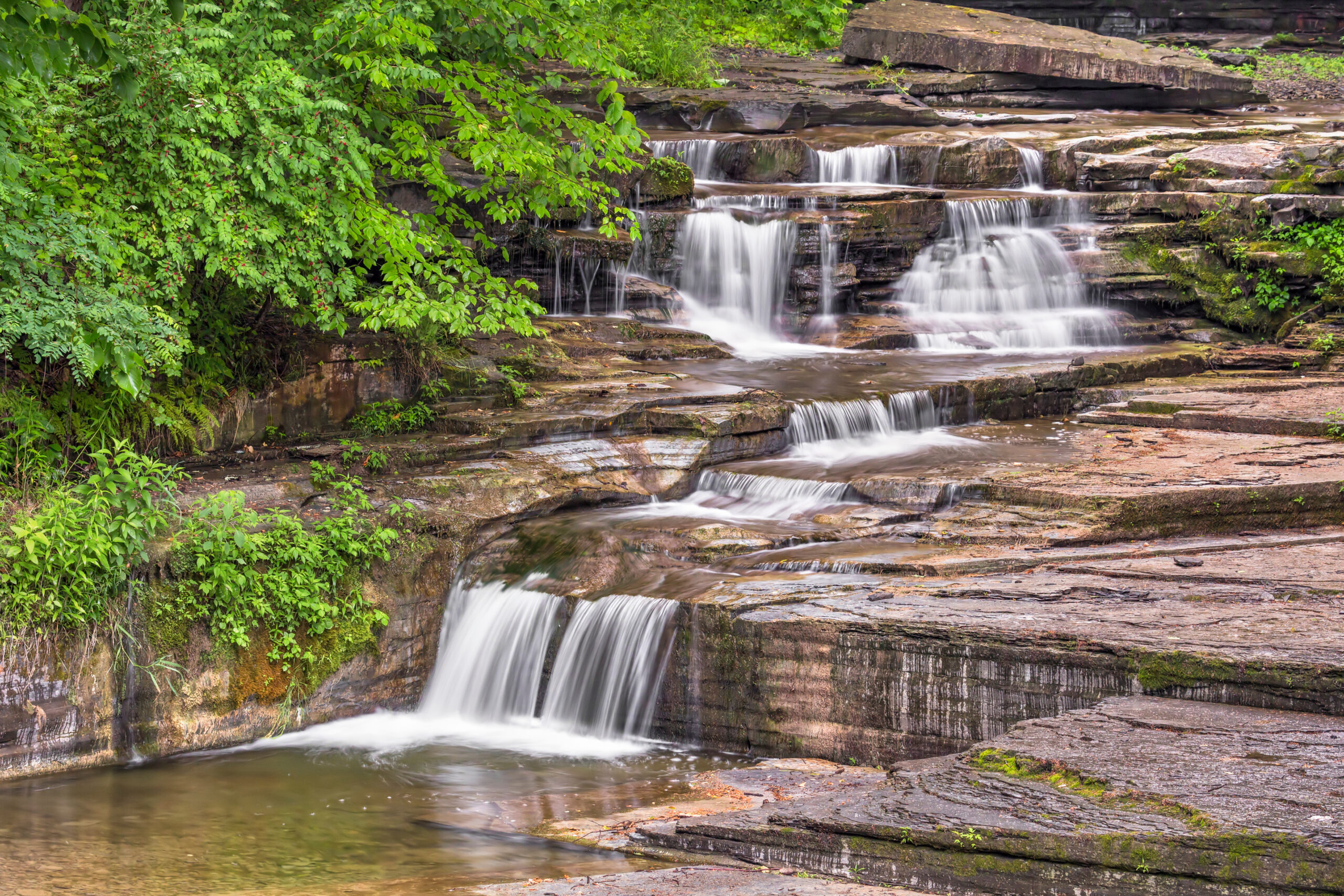 A serene waterfall cascades over layered rocks, surrounded by lush green vegetation at Havana Glen. The water flows gently, creating a series of small, tranquil pools in this natural setting near Havana Glen Trailer Park.