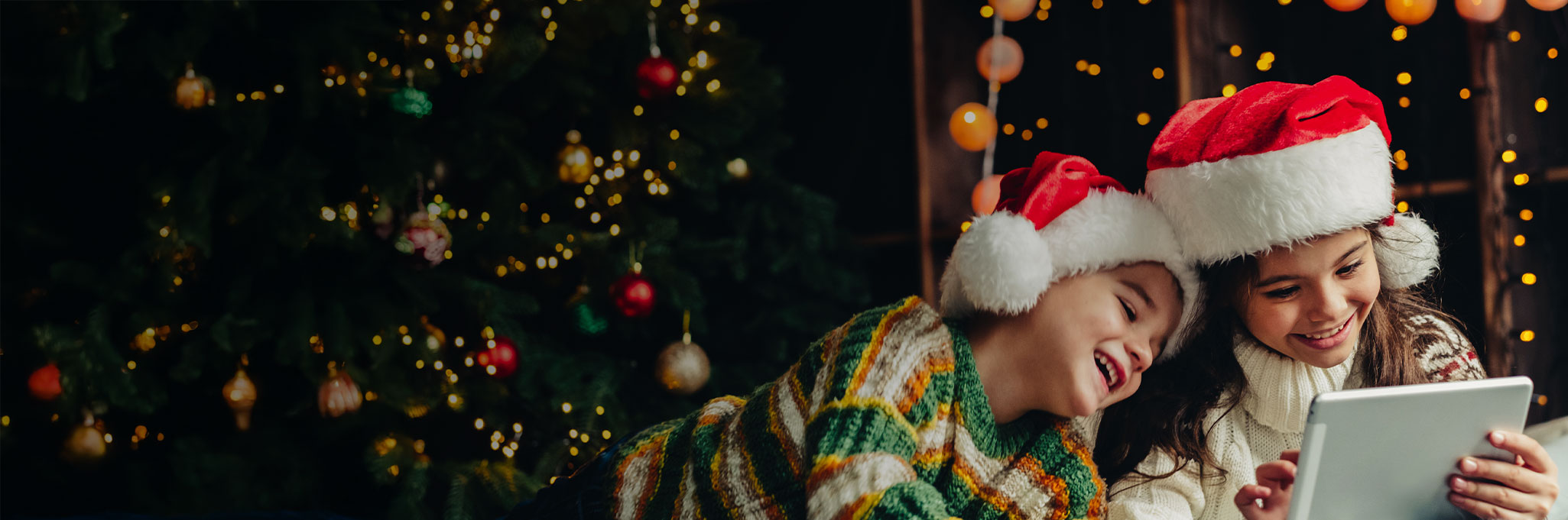 Two children wearing Santa hats laugh while looking at a tablet connected to high-speed internet, surrounded by warm Christmas lights and a decorated tree.